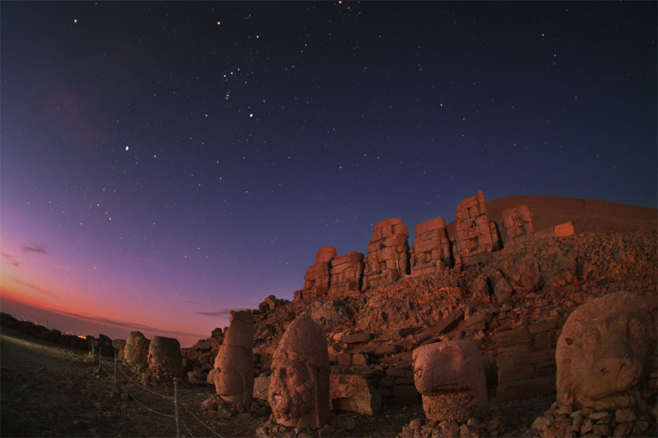 Een leeuw, een vogel en enkele mensenhoofden met hoge hoofddeksels (of verlengde schedels?) zien de zon opkomen bij de nog niet blootgelegde piramide van Nemrut Dağı in Turkije. Hoog aan de sterrenhemel houden de ster Sirius en het sterrenbeeld Orion, wellicht het thuis van hun makers, nog altijd de wacht over hen
